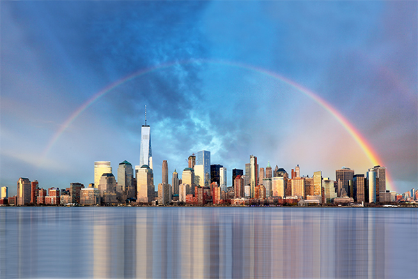 photo of New York City skyline with water in the foreground and a rainbow in the blue sky above the buildings