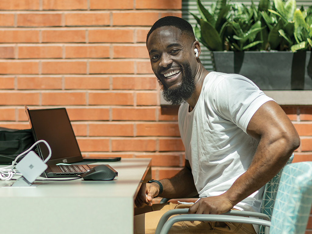 A man smiles seated at a desk with two laptops open in front of him.